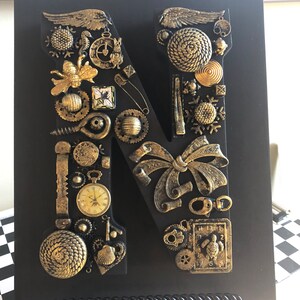 Peut inclure: Une lettre "N" en bois noir décorée de divers ornements dorés de style steampunk. Les décorations comprennent des engrenages, des ailes, une montre de poche et un nœud. La lettre est affichée sur un fond noir.