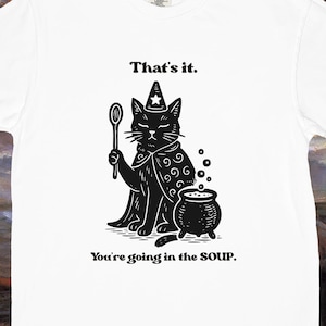 May include: White t-shirt featuring a black illustration of a cat in a wizard's hat and cape, holding a spoon, with a cauldron. Text reads "That's it. You're going in the SOUP."