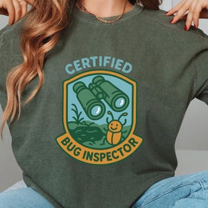 Peut inclure: T-shirt vert olive avec un motif "CERTIFIED BUG INSPECTOR". Le motif présente des jumelles, un insecte de dessin animé souriant et le texte en bleu et jaune. Le t-shirt est en matière souple.