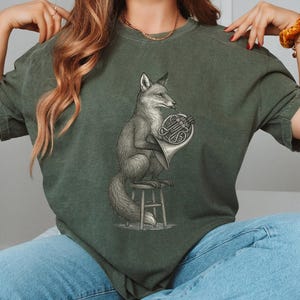 Könnte beinhalten: Grünes T-Shirt mit einer detaillierten Illustration eines Fuchses, der ein Waldhorn spielt. Der Fuchs sitzt auf einem kleinen Hocker, dargestellt in einem Vintage-Gravurstil. Das T-Shirt hat eine einfarbige grüne Farbe.