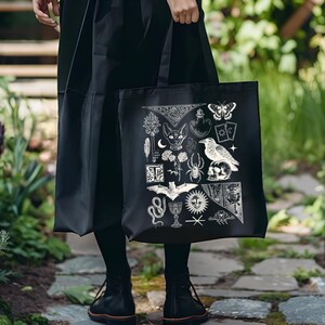 May include: Black tote bag featuring a white illustration of various occult symbols, including a cat, raven, spider, and butterfly. The bag is held by a person wearing a black outfit, standing on a stone path surrounded by greenery.