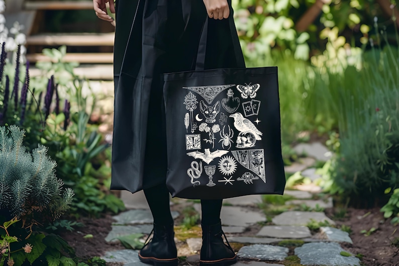May include: Black tote bag featuring a white illustration of various occult symbols, including a cat, raven, spider, and butterfly. The bag is held by a person wearing a black outfit, standing on a stone path surrounded by greenery.