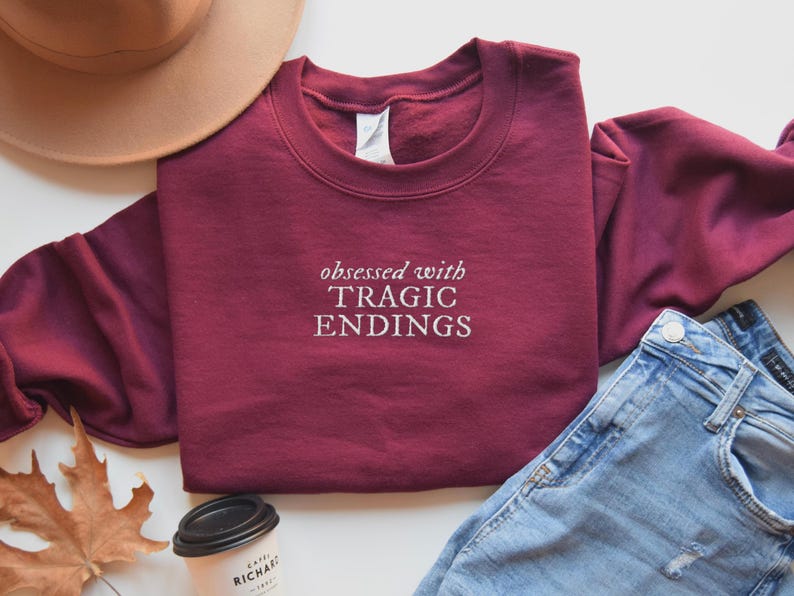 a casual outfit consisting of a burgundy sweatshirt with the text "obsessed with TRagic Endings" printed on it, a beige hat, a pair of blue jeans, and a coffee cup.