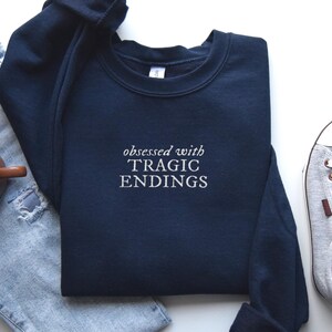A navy blue sweatshirt with the text "obsessed with TRagic Endings" printed on it, along with a pair of jeans, a pair of sneakers, and a cup of coffee.