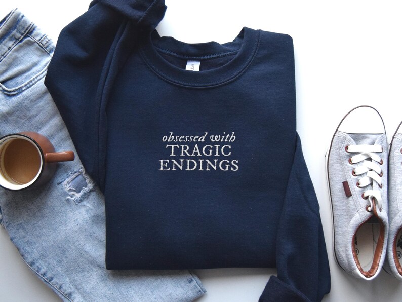 A navy blue sweatshirt with the text "obsessed with TRagic Endings" printed on it, along with a pair of jeans, a pair of sneakers, and a cup of coffee.