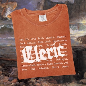 May include: Burnt orange t-shirt with the word "Cleric" in a stylized font. The shirt features a list of phrases related to role-playing games. The shirt is folded and has a tag in the upper right corner.
