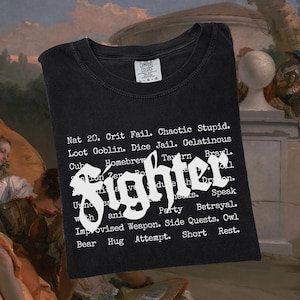 May include: Black t-shirt with the word "Fighter" in white, stylized font. The shirt features a list of phrases and words in white, including "Nat 20," "Crit Fail," and "Chaotic Stupid."
