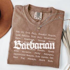 May include: A brown t-shirt with the word "Barbarian" in a gothic font, and a list of related terms. A silver necklace with a pendant is draped across the shirt. A brown hat and dried leaves are in the background.
