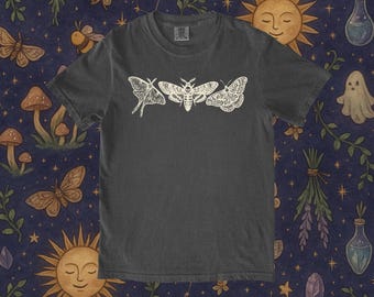 May include: Dark gray t-shirt featuring a white graphic of three moths. The design is centered on the shirt and is a detailed illustration. The shirt is made of a soft material and is suitable for casual wear.