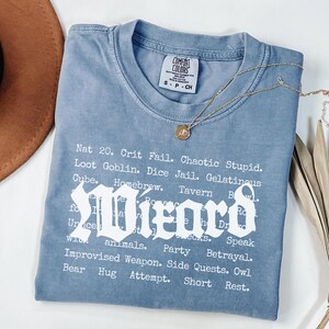 May include: A blue Comfort Colors t-shirt with white text listing phrases like "Nat 20" and "Wizard". A gold necklace with a pendant rests on the shirt. The shirt is folded and displayed with a brown hat and dried plant.