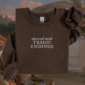 a brown sweatshirt with the text "obsessed with TRagic Endings" printed on it.