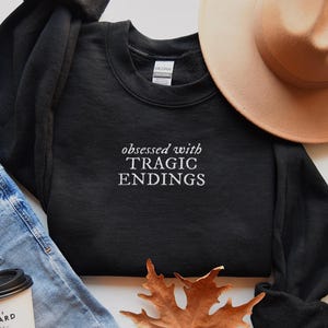 a casual outfit consisting of a black sweatshirt with the text "obsessed with TRagic Endings" printed on it, a beige hat, blue jeans, and a coffee cup.