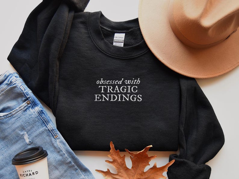 a casual outfit consisting of a black sweatshirt with the text "obsessed with TRagic Endings" printed on it, a beige hat, blue jeans, and a coffee cup.