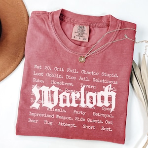 May include: A red t-shirt with the word "Warlock" in a gothic font. The shirt has a list of words below the main text, including "Nat 20", "Loot Goblin", and "Party". A gold necklace with a pendant is on the shirt.