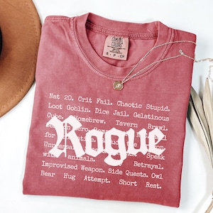 May include: A red t-shirt with the word "Rogue" in large white letters. The shirt has a list of words and phrases related to role-playing games. A gold necklace with a pendant rests on the shirt.