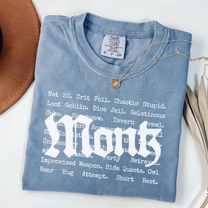 May include: A blue Comfort Colors t-shirt with the word "Monk" in a gothic font. The shirt features a list of phrases and terms related to role-playing games. A gold necklace with a pendant rests on the shirt.