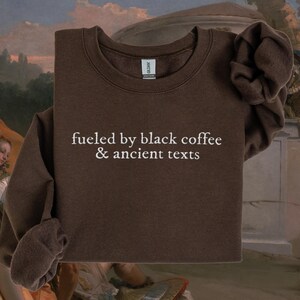 A brown sweatshirt with the text "fueled by black coffee & ancient texts" printed on it, displayed against a colorful background with a painting of a woman in the background.