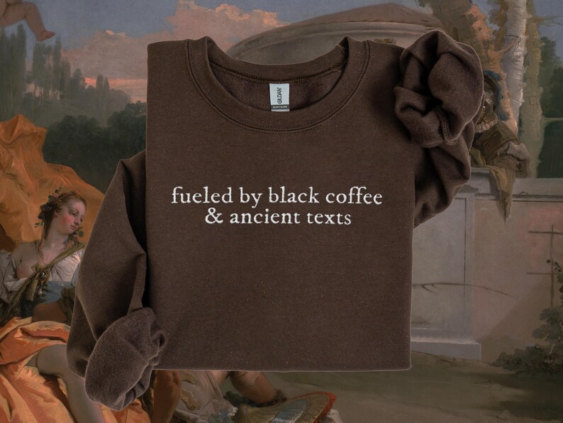 A brown sweatshirt with the text "fueled by black coffee & ancient texts" printed on it, displayed against a colorful background with a painting of a woman in the background.