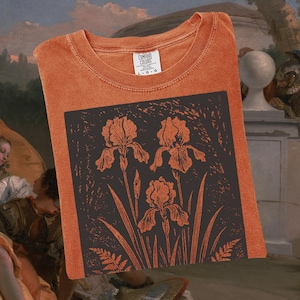May include: Orange t-shirt with a dark square graphic featuring three iris flowers and foliage. The shirt has a crew neck and a tag on the collar. The shirt is folded.