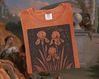 Puede incluir: Camiseta naranja con un diseño gráfico cuadrado oscuro con tres flores de iris y follaje. La camiseta tiene cuello redondo y una etiqueta en el cuello. La camiseta está doblada.