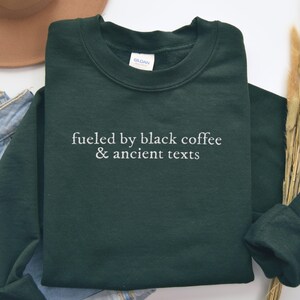 A green sweatshirt with the text "fueled by black coffee & ancient texts" printed on it, along with a pair of jeans and a hat.