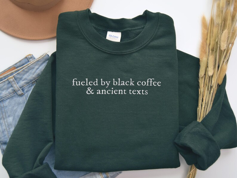 A green sweatshirt with the text "fueled by black coffee & ancient texts" printed on it, along with a pair of jeans and a hat.