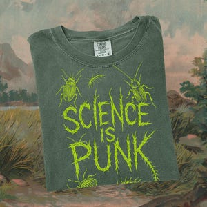 May include: A dark green t-shirt with the text "SCIENCE IS PUNK" in bright green, along with insect illustrations. The shirt is a Comfort Colors brand, and the design is screen printed. The shirt is folded and displayed on a painted background.