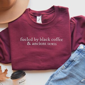 A burgundy sweatshirt with the text "fueled by black coffee & ancient texts" printed on it, along with a beige hat, a pair of jeans, and a coffee cup.