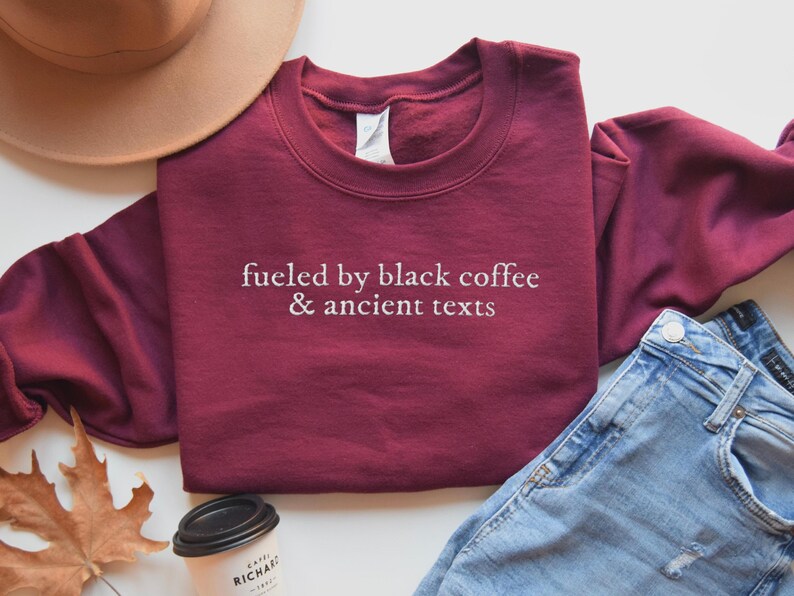 A burgundy sweatshirt with the text "fueled by black coffee & ancient texts" printed on it, along with a beige hat, a pair of jeans, and a coffee cup.
