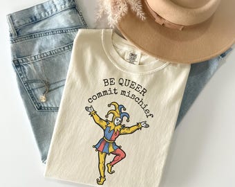Puede incluir: Camiseta color crema con el texto "BE QUEER commit mischief" y un dibujo de un bufón de dibujos animados en amarillo, azul y rojo. La camiseta se combina con vaqueros de mezclilla de lavado claro, un sombrero beige y un collar dorado.