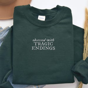 A dark green sweatshirt with the text "obsessed with TRagic Endings" printed on it, along with a pair of jeans and a hat.