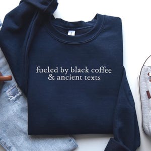 A navy blue sweatshirt with the text "fueled by black coffee & ancient texts" printed on it, along with a pair of jeans, a pair of sneakers, and a cup of coffee.
