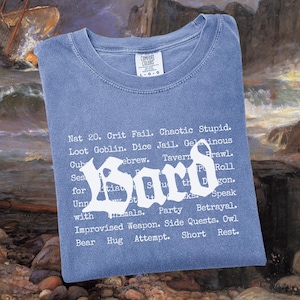 May include: A blue t-shirt with the word "Bard" in a gothic font, surrounded by a list of fantasy role-playing terms. The shirt is made of a soft material and is perfect for fans of tabletop games.