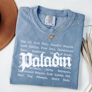 May include: A blue Comfort Colors t-shirt with the word "Paladin" in a gothic font. The shirt features a list of phrases and words. A gold necklace with a pendant is draped across the shirt.