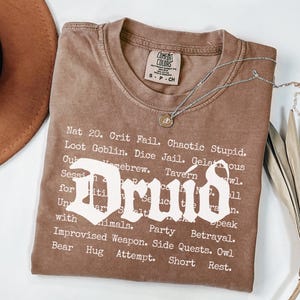 May include: A brown t-shirt with the word "Druid" in a bold, white font. The shirt features a list of phrases and terms related to role-playing games. A silver necklace with a pendant is draped across the shirt.
