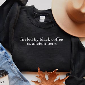 a casual outfit consisting of a black sweatshirt with the text "fueled by black coffee & ancient texts" printed on it, a beige hat, a pair of blue jeans, and a coffee cup.