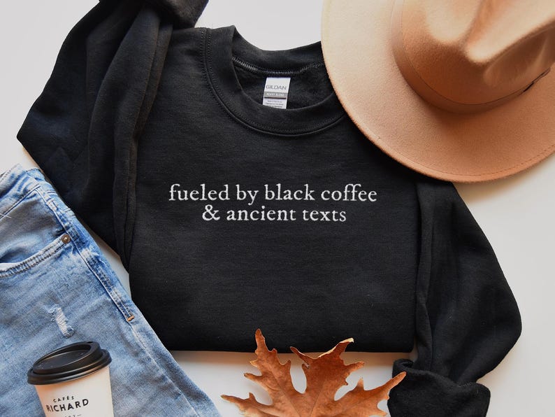 a casual outfit consisting of a black sweatshirt with the text "fueled by black coffee & ancient texts" printed on it, a beige hat, a pair of blue jeans, and a coffee cup.
