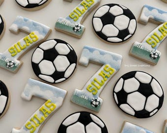 Puede incluir: Un conjunto de galletas de azúcar decoradas con temática de fútbol. Las galletas están decoradas con balones de fútbol blancos y negros, hierba verde, nubes azules y el nombre "Silas" en glaseado amarillo.