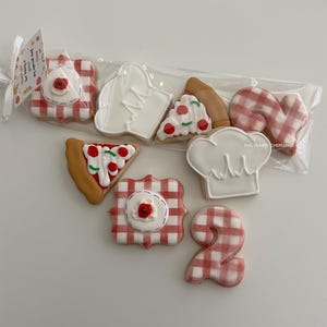 May include: A collection of decorated sugar cookies in a clear plastic bag, featuring shapes of pizza slices, chef hats, the number two, and square plaques with red and white gingham patterns. A small tag attached reads: Thank you for being a pizza my special day - Drew.