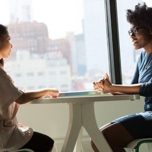 May include: Two people sitting at a table in front of a window. The person on the left is wearing a light brown shirt and the person on the right is wearing a blue shirt and glasses.