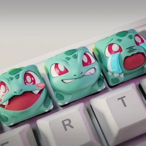May include: Three custom keycaps featuring Bulbasaur from Pokémon with different facial expressions. The keycaps are green with red eyes and are on a white keyboard. The Nom Nom Figures logo is in the top left corner.