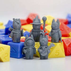 Puede incluir: Un conjunto de figuras grises con forma de gato, cada una vestida con atuendos de fantasía, incluyendo un mago, un arquero y un bardo. Las figuras se colocan sobre un fondo de dados coloridos, sugiriendo un tema de juego de mesa.