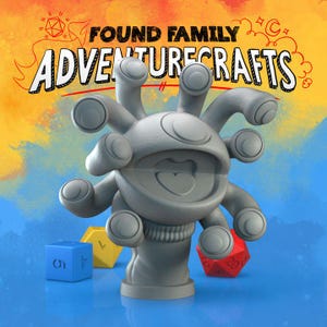 May include: A gray, stylized monster figurine with multiple eye stalks and a wide grin. The image includes the text "FOUND FAMILY ADVENTURECRAFTS" in a playful font. Colorful dice are scattered around the base of the figurine, suggesting a tabletop game theme.