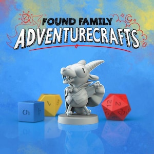May include: A gray dragon figurine with a cape and horns stands on a base, next to a blue six-sided die, a yellow eight-sided die, and a red twenty-sided die. The text "FOUND FAMILY ADVENTURECRAFTS" is above the dragon.