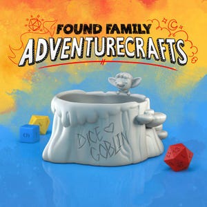 May include: A white ceramic dice holder shaped like a tree stump, with a small goblin figure perched on top. The holder has the words "Dice Goblin" written on it. The image also includes several dice and the text "Found Family Adventurecrafts".