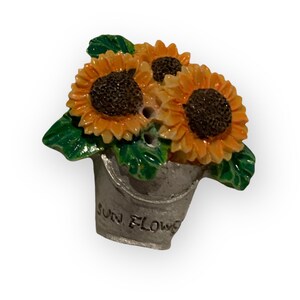 May include: A small, white ceramic bucket with three yellow sunflowers with brown centres. The bucket has the words "SUN FLOWER" written on the side.