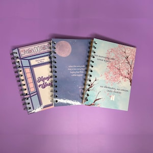 Könnte beinhalten: Drei spiralgebundene Notizbücher mit unterschiedlichen Designs. Das erste Notizbuch hat eine blaue und rosafarbene Ladenfront mit dem Text "Willkommen im Magic Shop". Das zweite Notizbuch hat einen blauen und violetten Himmel mit einem Vollmond und Text, der lautet "Wenn dieses Lied endet, kann ein neues Lied beginnen, in der Hoffnung, dass ich ein bisschen glücklicher sein werde". Das dritte Notizbuch hat einen blauen und rosafarbenen Himmel mit einem rosafarbenen Kirschblütenbaum und Text, der lautet "Der Morgen wird wiederkommen, keine Dunkelheit, keine Jahreszeit dauert ewig".