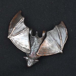 May include: A silver and copper bat pendant with detailed wings. The bat is in a flying position with its wings spread out.