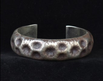Puede incluir: Una pulsera de plata con una superficie texturizada y martillada. La pulsera tiene una banda ancha con una serie de hendiduras circulares. El metal tiene un aspecto ligeramente oxidado, con tonos más oscuros en las zonas hundidas. Sobre fondo negro.