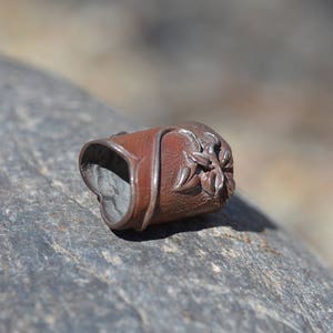 Op de afbeelding: Een kleine, bruine leren vingerbeschermer met een bloemmotief. De beschermer is gemaakt van leer en heeft een metalen ring aan de achterkant.